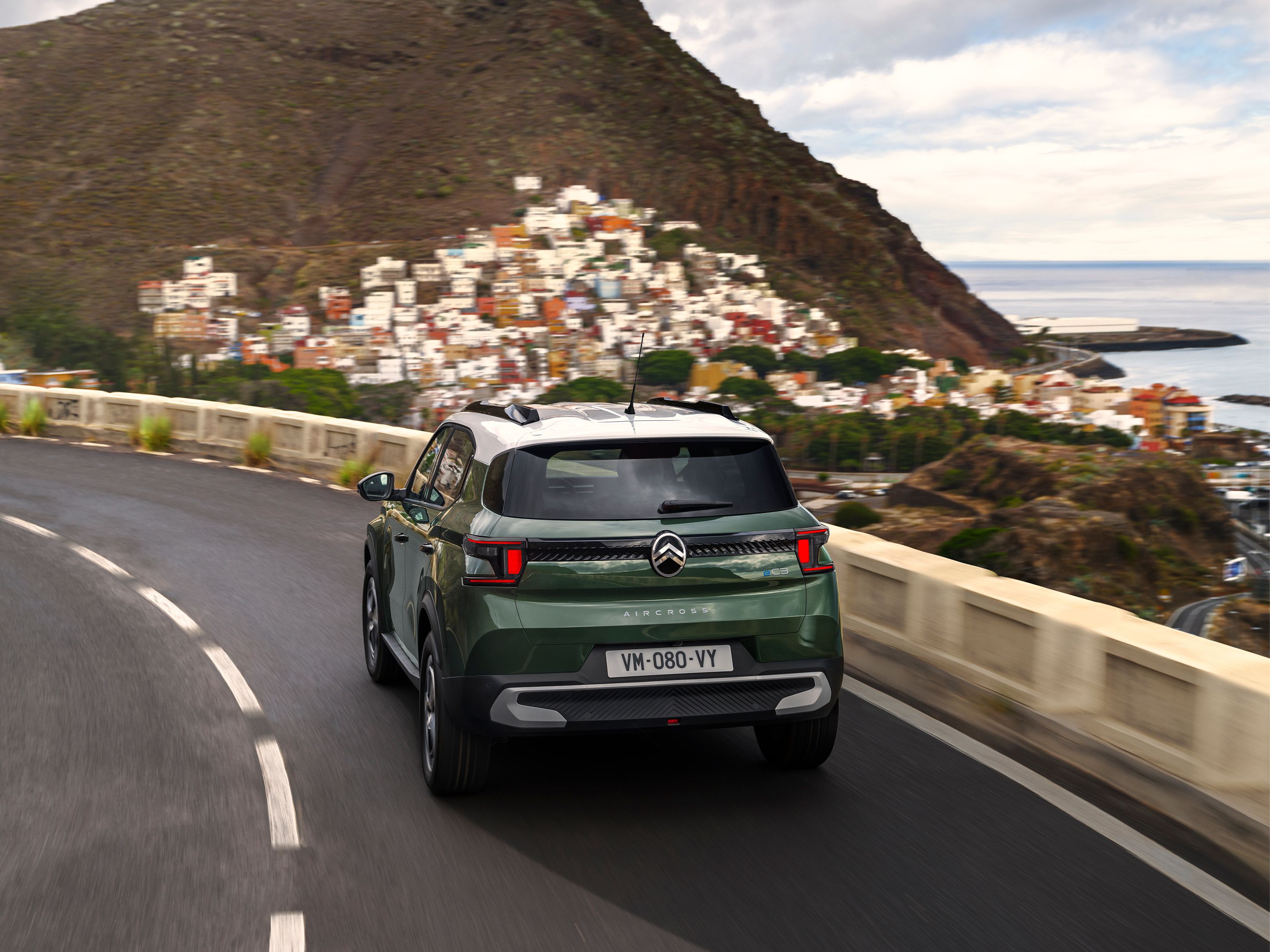 Rear three‑quarter driving view of Citroën ë-C3 Aircross SUV on a coastal road for confident, smooth and affordable everyday mobility.